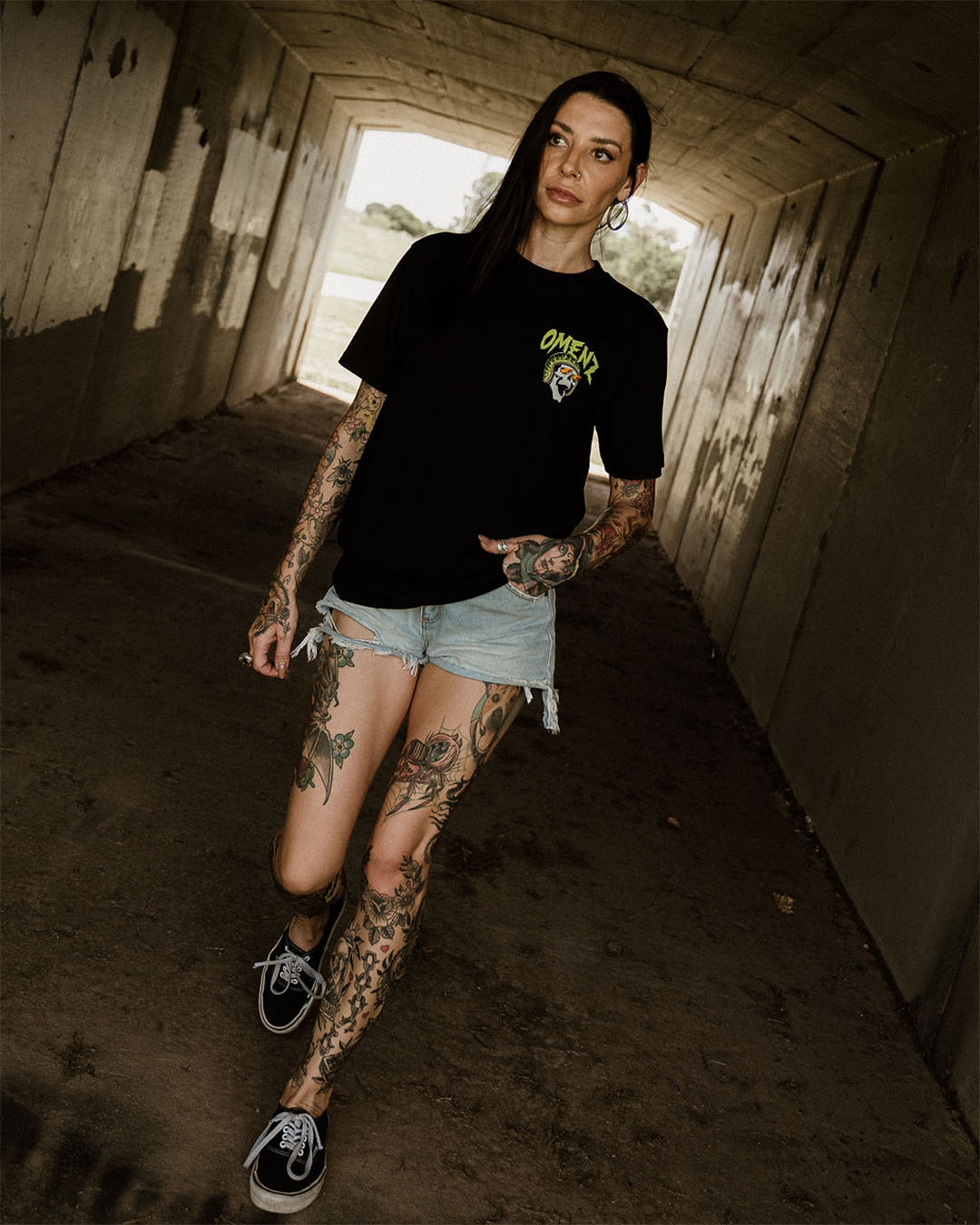 Tattooed female wearing denim shorts and an Omenz Killswitch T-shirt walking through an urban underpass