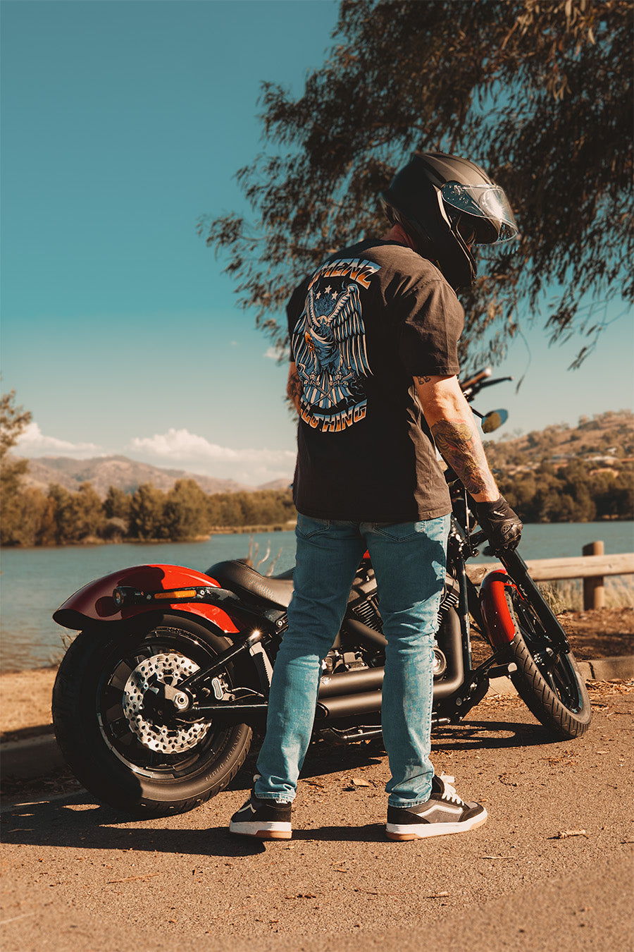 Man wearing an Omenz oversized graphic tee t-shirt and a motorcycle helmet standing next to a Harley Davidson at a lake