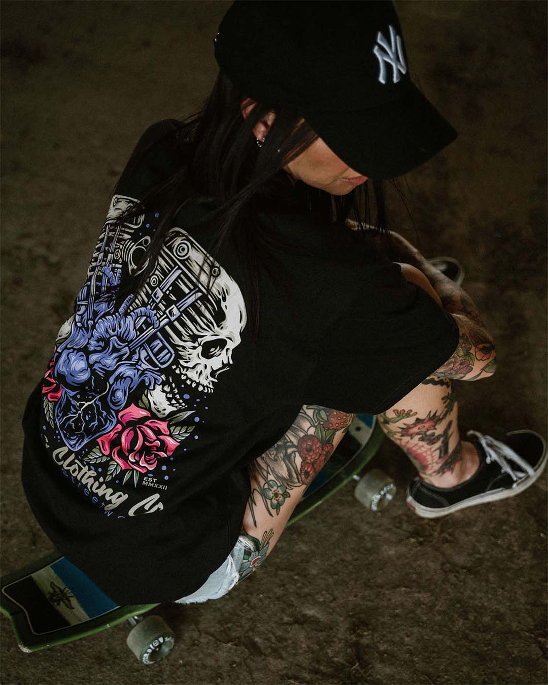 Tattooed female wearing an Omenz Full Throttle T-shirt, baseball cap and Van shoes sitting on a skateboard