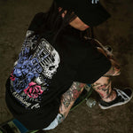 Tattooed female wearing an Omenz Full Throttle T-shirt, baseball cap and Van shoes sitting on a skateboard