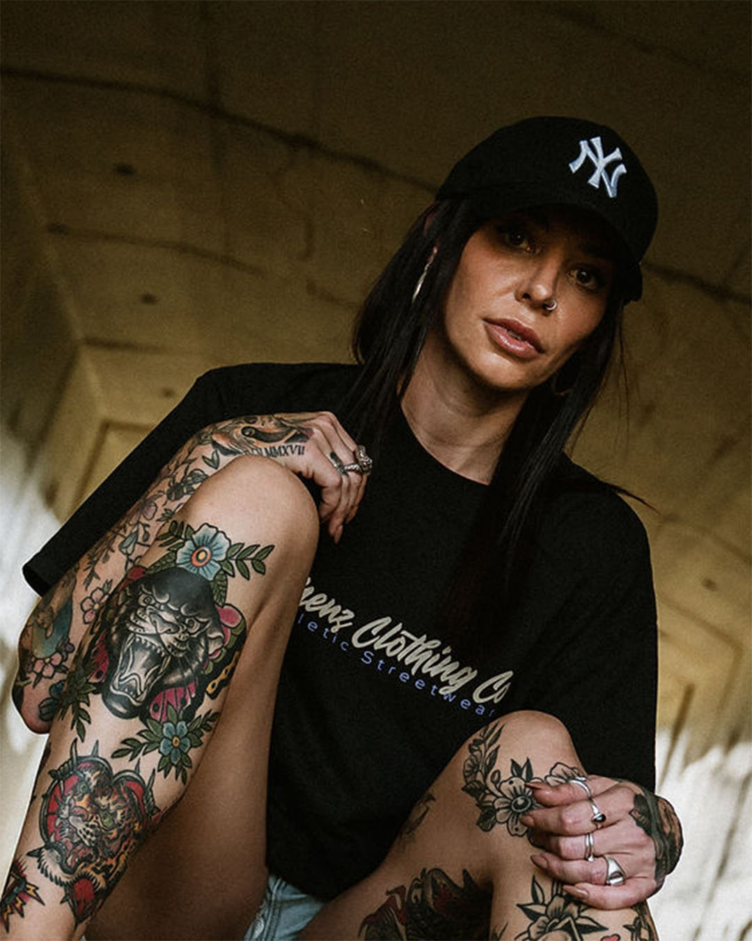 Tattooed female wearing an Omenz Full Throttle T-shirt and NY baseball cap in an urban underpass