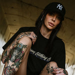 Tattooed female wearing an Omenz Full Throttle T-shirt and NY baseball cap in an urban underpass