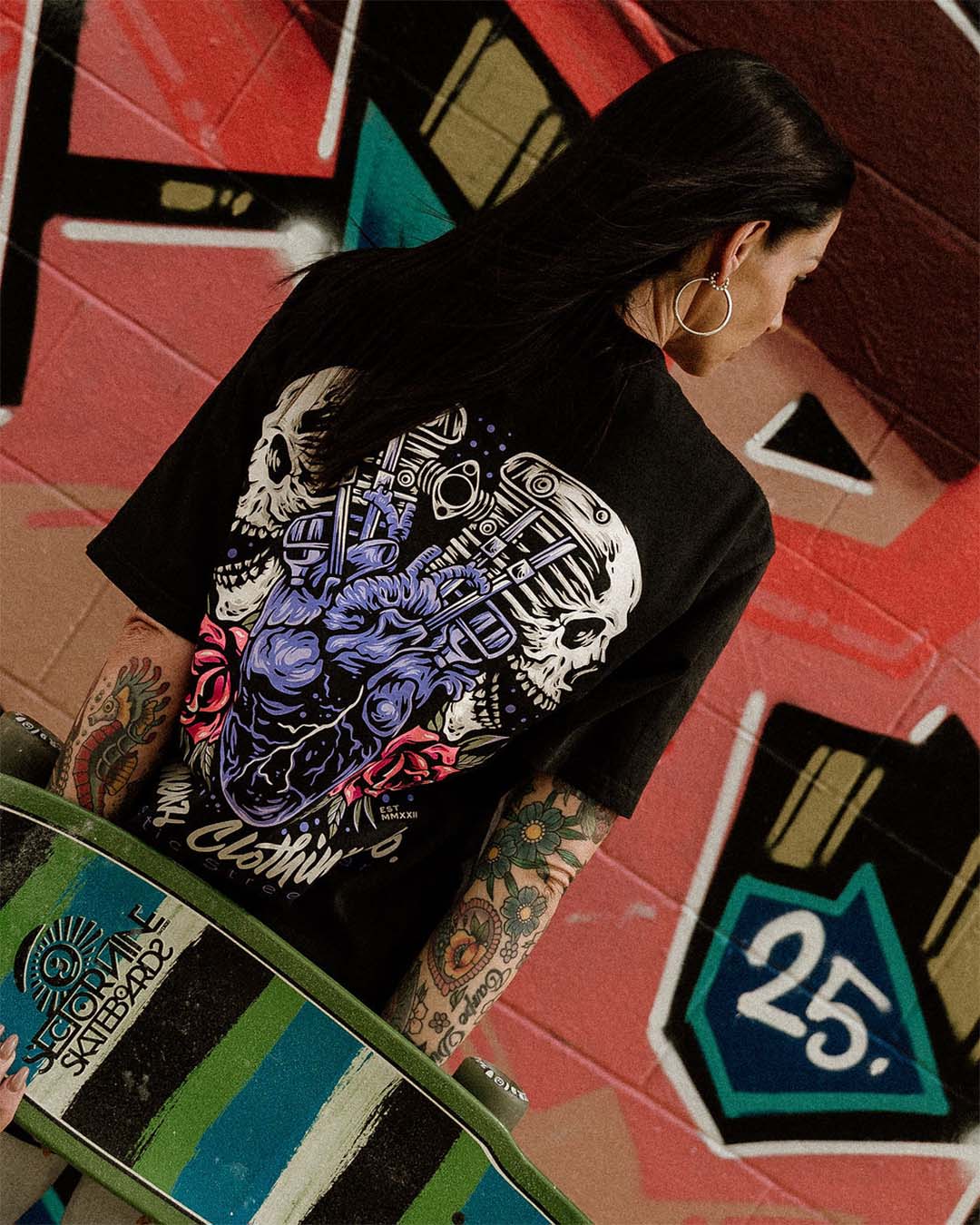 Tattooed female wearing an Omenz Full Throttle T-shirt and a skateboard in an urban graffiti street