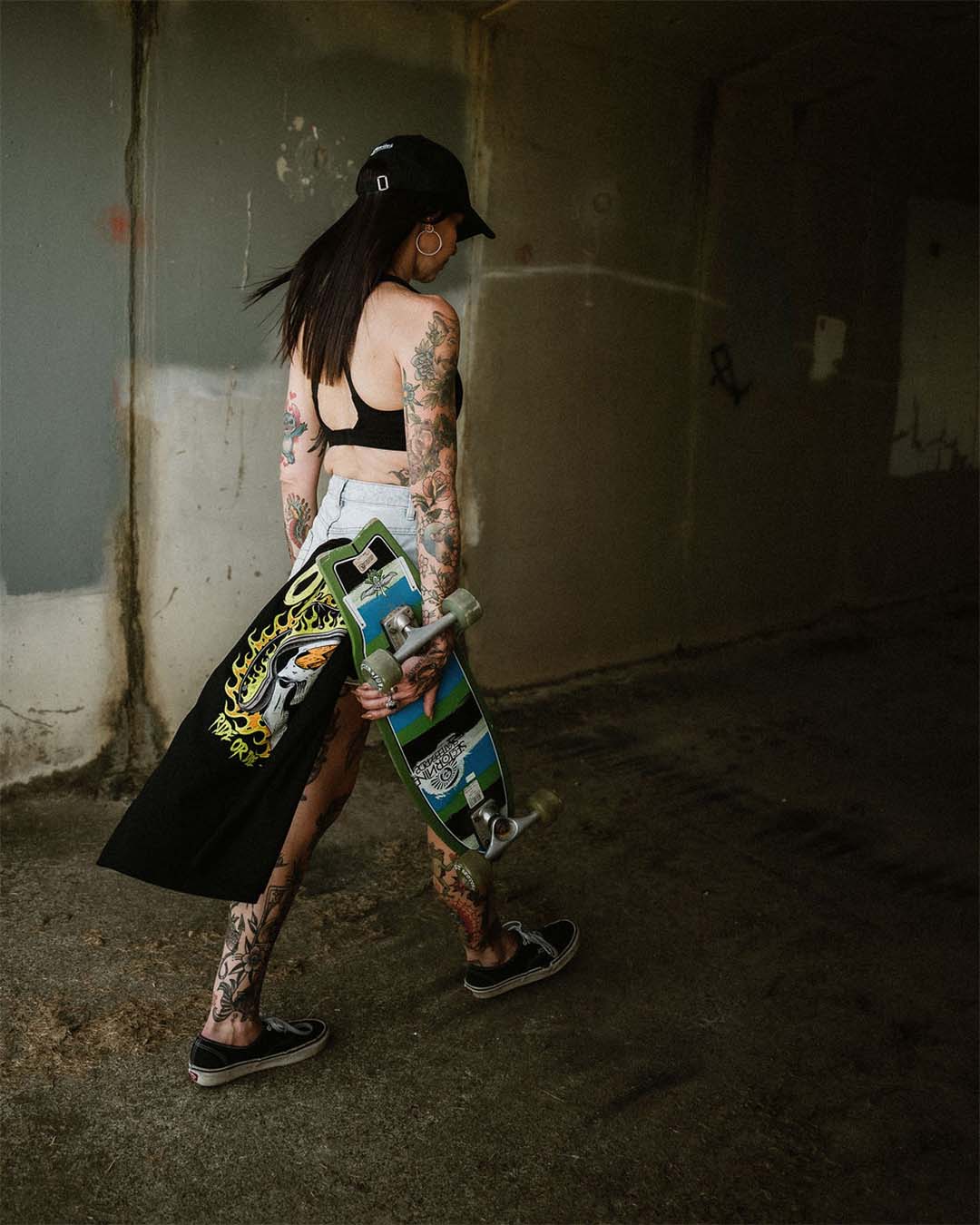 Tattooed female in denim shorts with an Omenz Killswitch T-shirt tucked into her jeans pocket. Woman is carrying a skateboard through an urban underpass